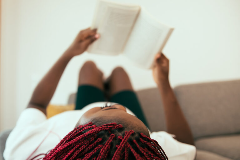 Black woman laying on the sofa reading a book with his feets on the wall